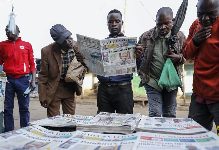 Un grupo de hombres se reúne alrededor de una selección de periódicos, uno de los cuales está leyendo uno.