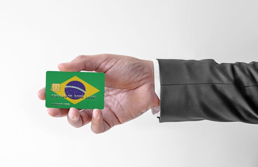 A man’s hand holds a plastic credit card decorated with the colors of the flag of Brazil.
