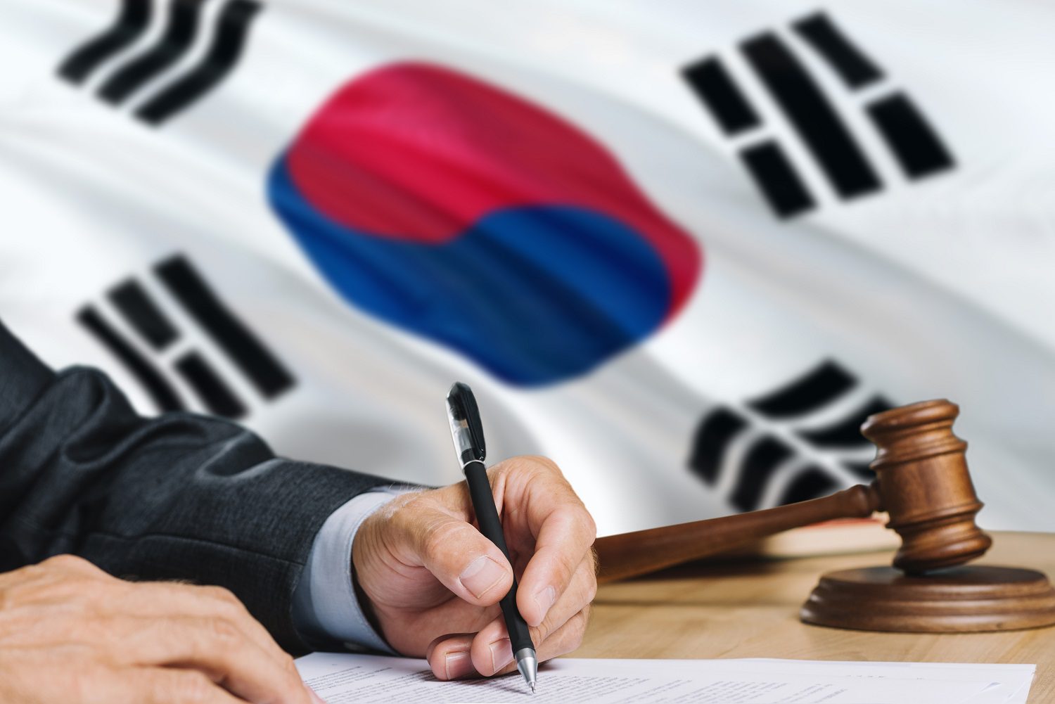 Una persona con traje de negocios escribe en un documento con la bandera de Corea del Sur y un martillo de madera y un tocón en el fondo.