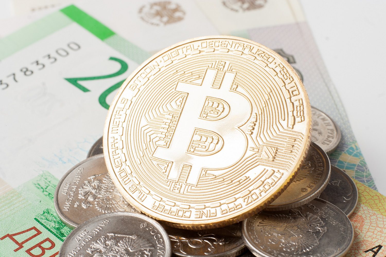 Una ficha de metal destinada a representar a Bitcoin se encuentra encima de una pila de monedas fiduciarias y billetes rusos.