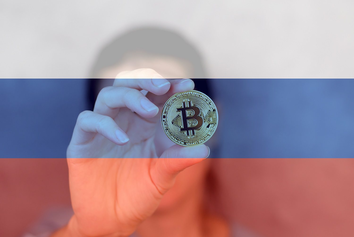 Una persona sostiene una ficha de metal destinada a representar Bitcoin contra el fondo de la bandera rusa.