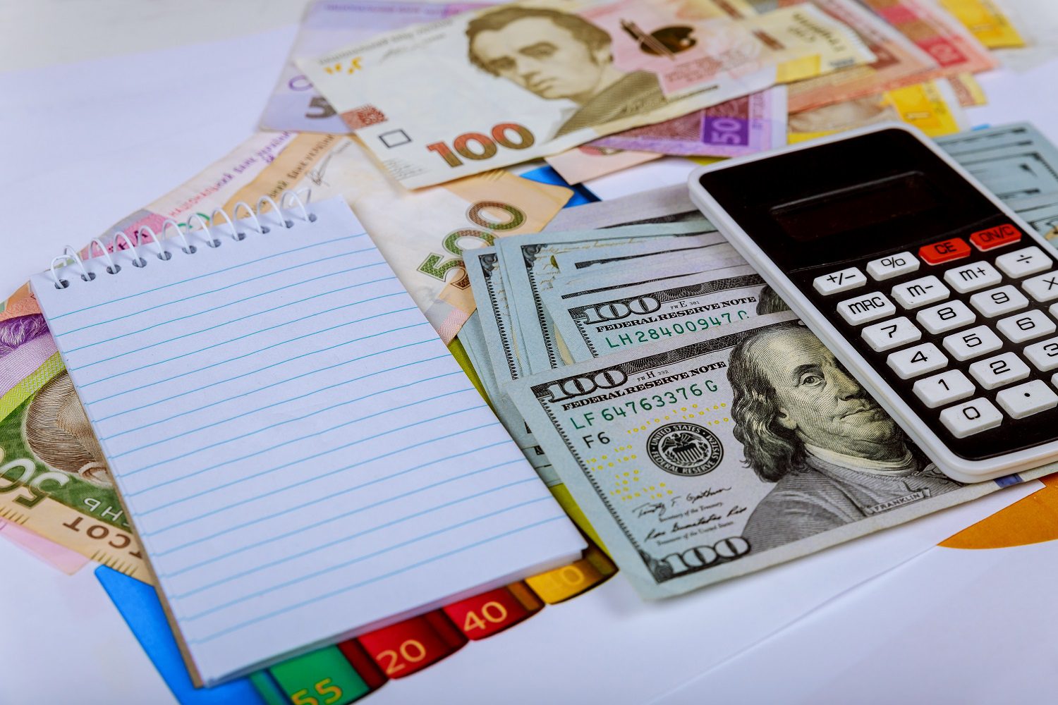 Una calculadora y un bloc de notas de papel descansan sobre una selección de billetes de USD y UAH.