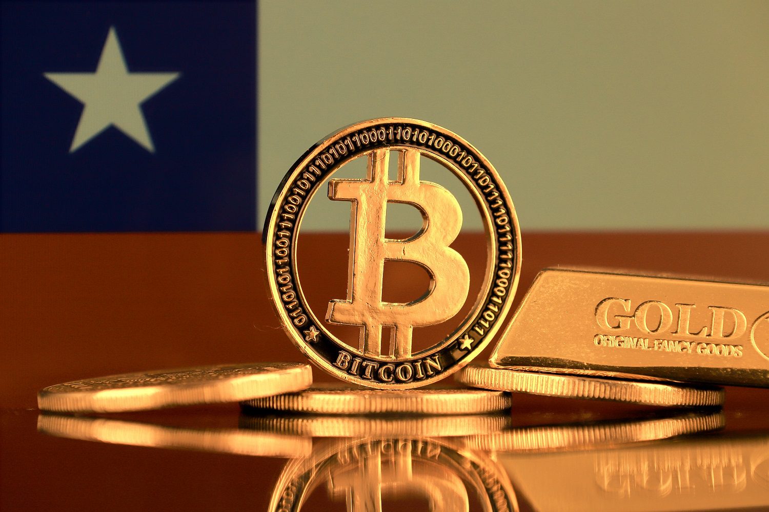 Un token de metal destinado a representar Bitcoin, junto con otros tokens, una barra de oro, con el telón de fondo de la bandera chilena.