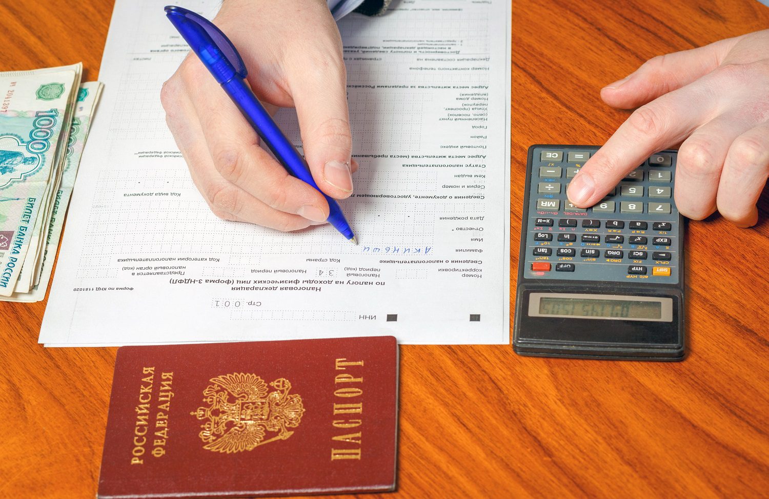 Una persona llena una declaración de impuestos rusa, sosteniendo un bolígrafo.  Al lado del formulario hay una pequeña pila de billetes rusos y una calculadora.  La persona presiona un botón de la calculadora.