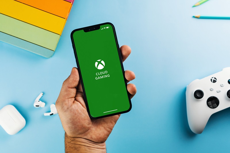 Man holding a smartphone with Xbox Cloud Gaming app on the screen. Blue background with school supplies, AirPods, video game controller. Rio de Janeiro, RJ, Brazil. April 2022